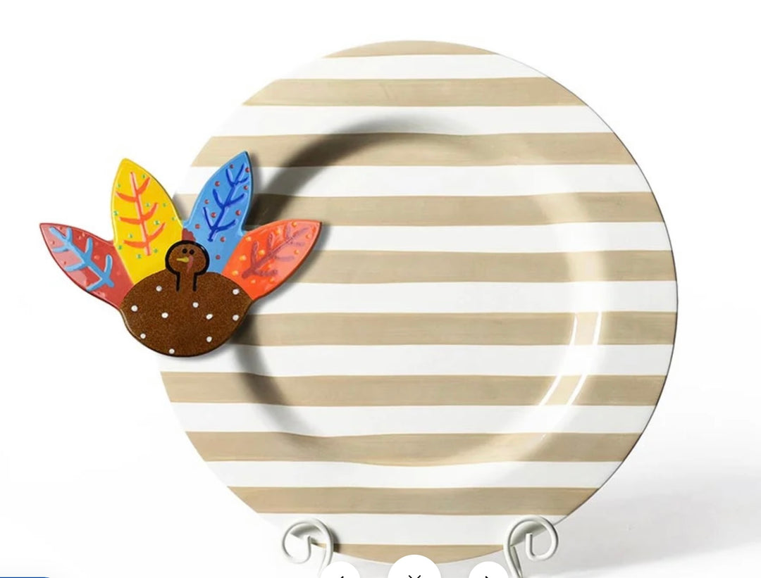 A round ceramic plate with horizontal beige and white stripes, decorated with a colorful wooden turkey magnet on the left side, displayed on a white stand against a white background.
