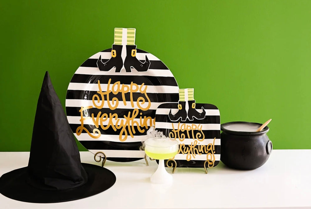 Halloween-themed table decor set including a black witch hat, striped platter with gold 'Happy Everything' lettering, a smaller matching sign, and a black cauldron, arranged on a white surface against a bright green background.
