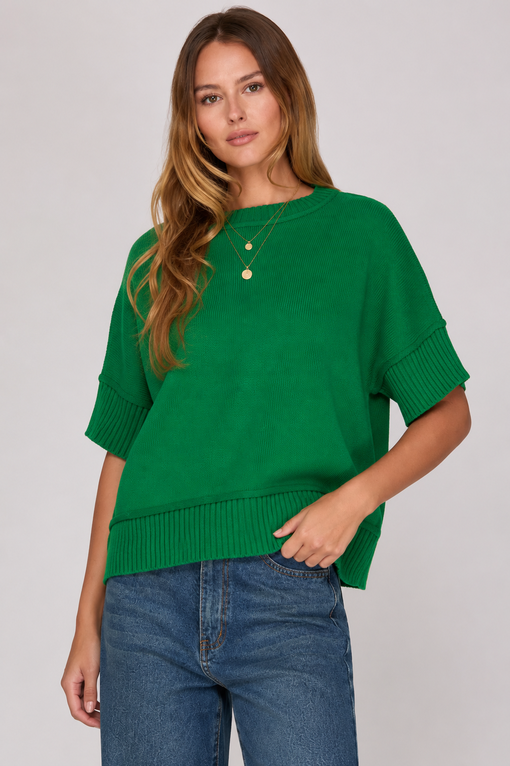 A woman wearing a bright green short-sleeve knit sweater with ribbed cuffs and hem, paired with blue jeans, against a light gray studio background.