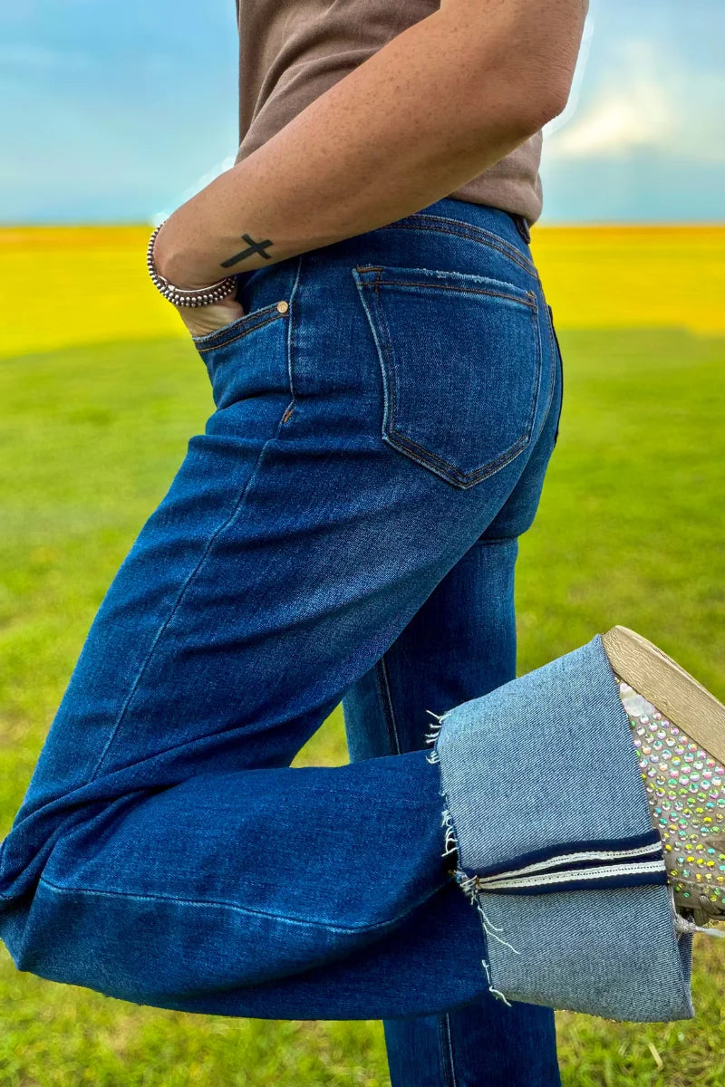 A person outdoors in a grassy field wearing dark blue wide-leg jeans with rolled cuffs, holding a woven straw bag. The photo is cropped from the chest down, showing the back pocket and side of the jeans.