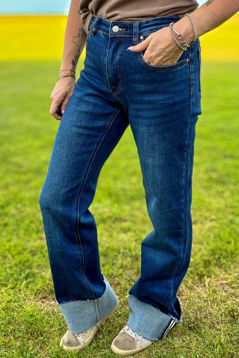 A person wearing dark blue cuffed jeans with a relaxed straight-to-wide leg fit, standing outdoors on a grassy field with a yellow field in the background.