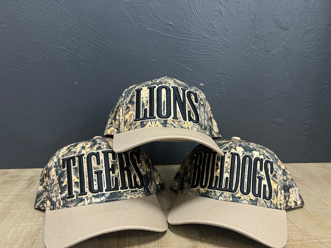 Stacked camo mascot hats featuring Lions, Tigers, and Bulldogs puff embroidery with light brown brims.