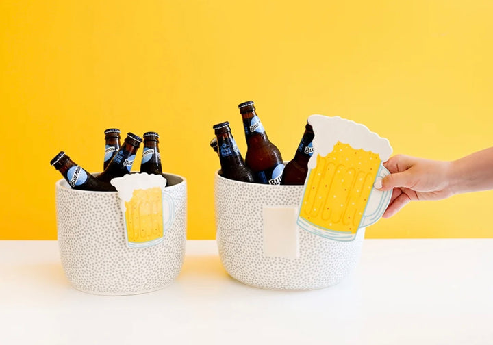 Two white speckled ceramic beer bucket coolers on a white table against a yellow background, filled with brown glass beer bottles, with a hand holding a beer mug cutout next to one bucket.