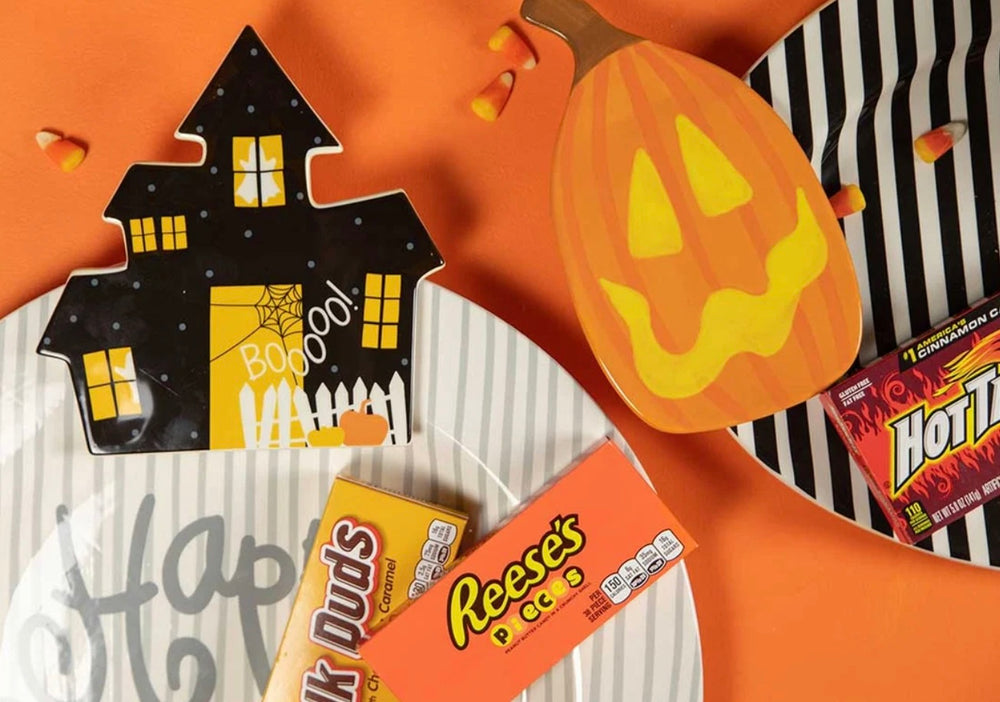 A Halloween-themed candy and snack arrangement on an orange background, featuring a jack-o'-lantern-shaped box, a house-shaped box, Reese's Pieces, Snickers Hots, and a bag of Reese's Dip.