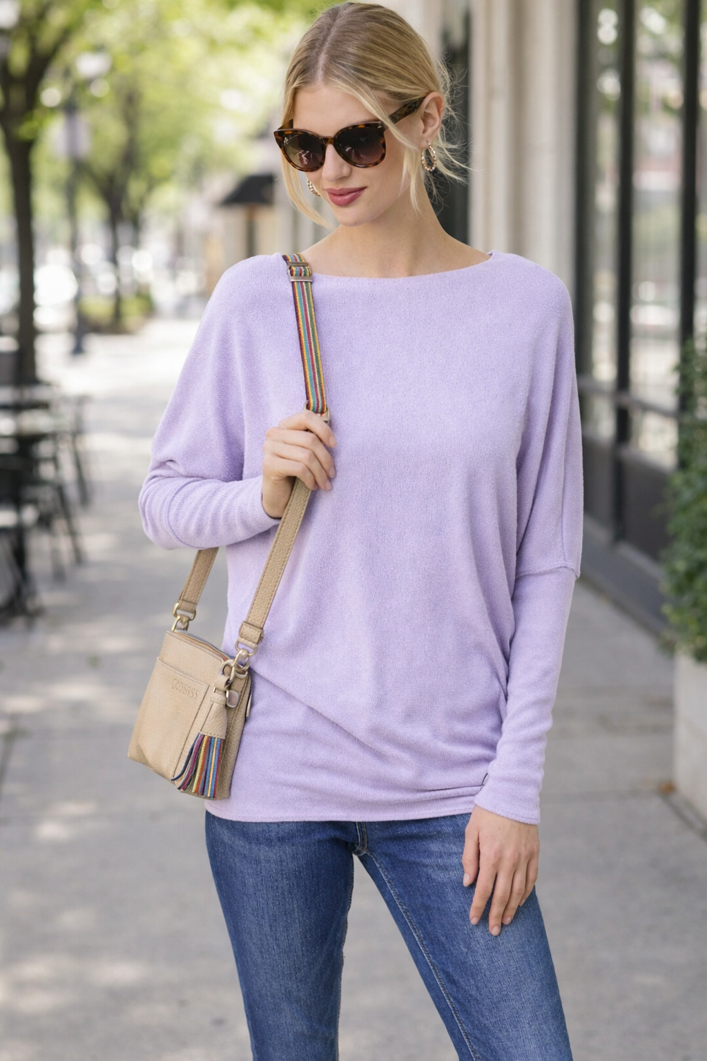 Woman wearing a lavender dolman sleeve top styled with jeans, sunglasses, and a crossbody bag walking on a sidewalk