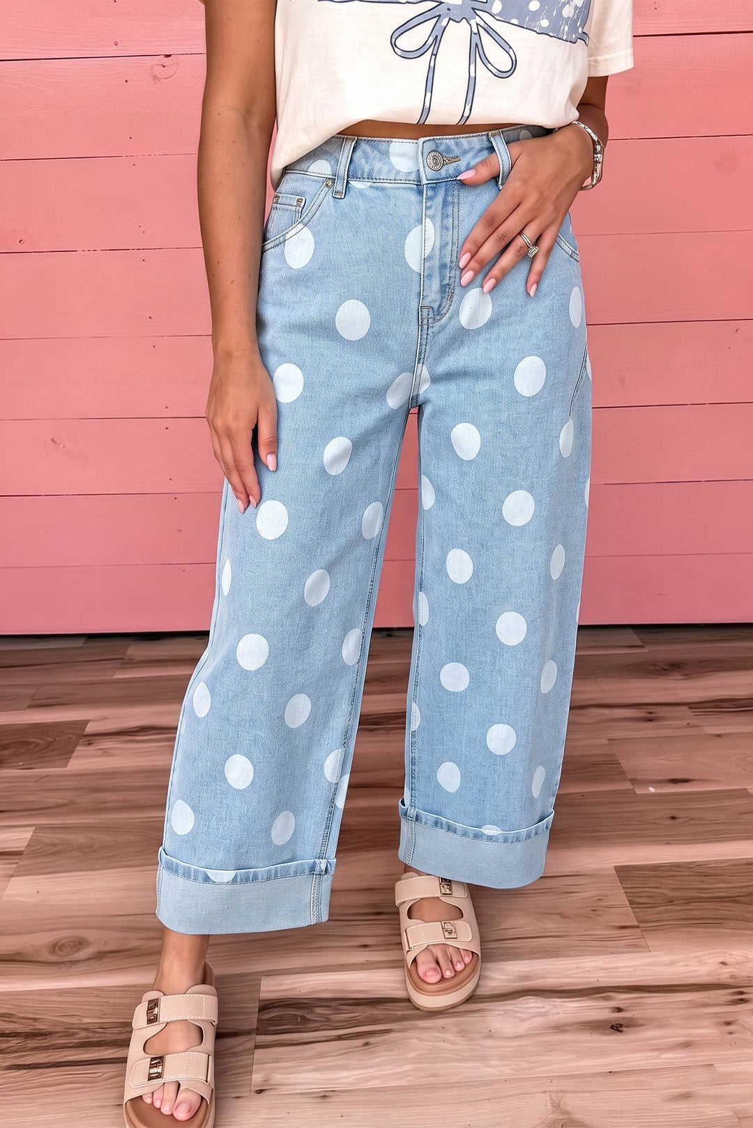 A person wearing light blue wide-leg denim jeans with large white polka dots, paired with a cream crop top and beige slide sandals, standing against a pink wood-paneled wall and wood floor.